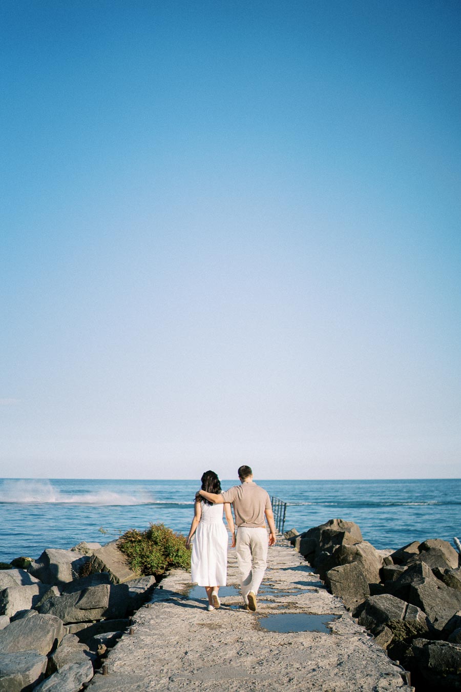 A couple walking arm in arm on a rocky jetty towards the ocean under a clear blue sky.