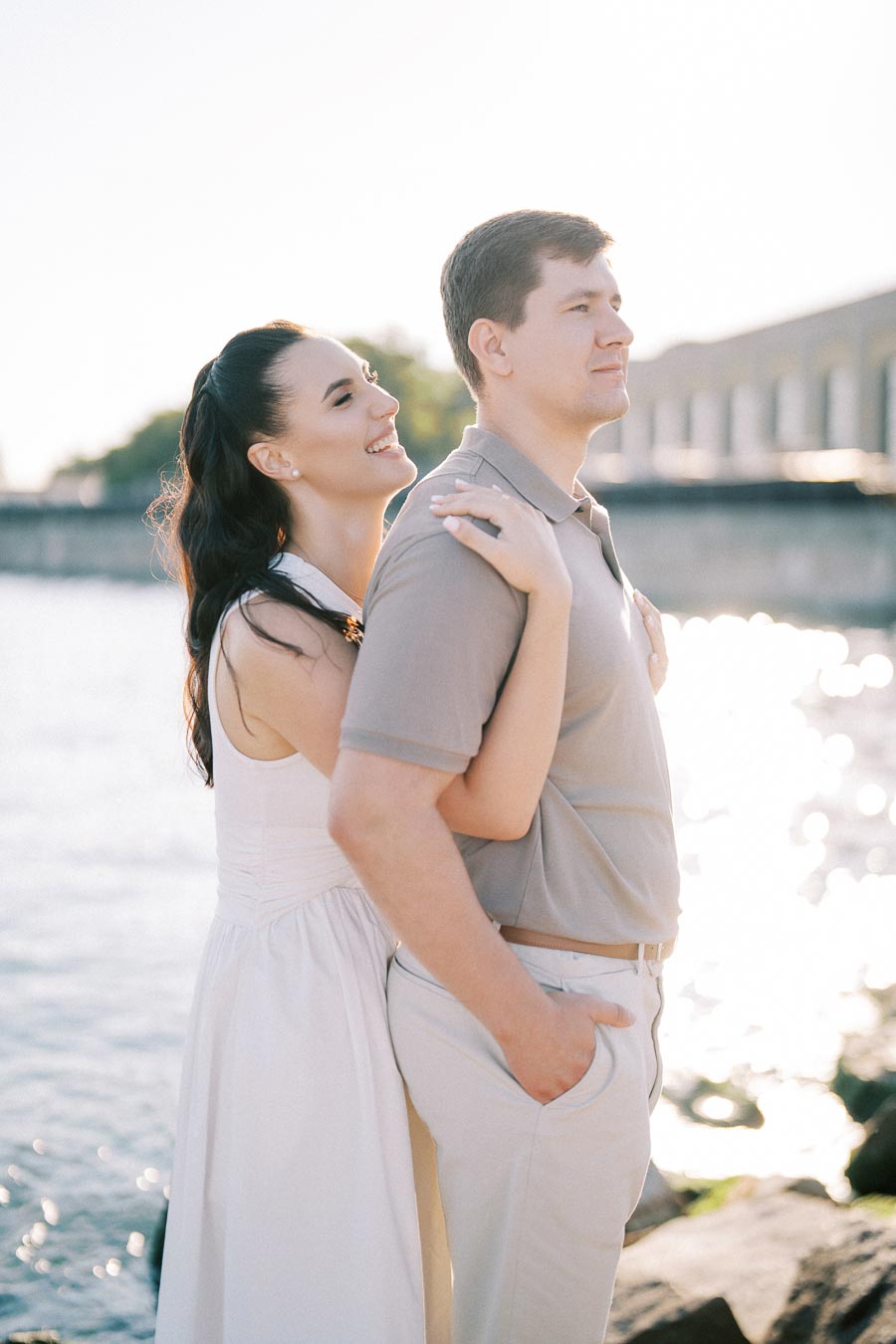 A happy couple enjoying a romantic moment by the waterfront, with sunlight reflecting off the water, the woman embracing the man from behind, both smiling contentedly.