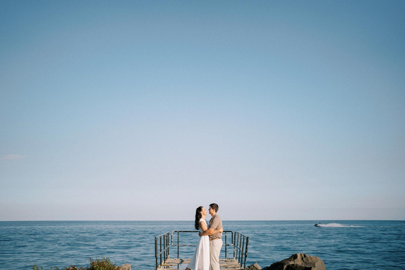 Couple embracing on an ocean pier under a clear blue sky, with a distant speedboat on the water.
