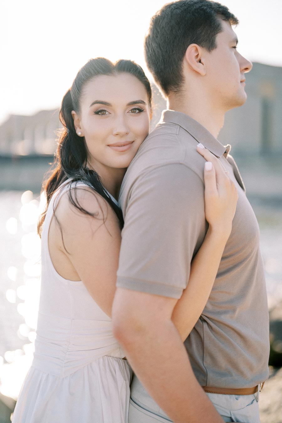Young woman embracing a man outdoors during a sunny day, with a serene waterfront setting in the background.