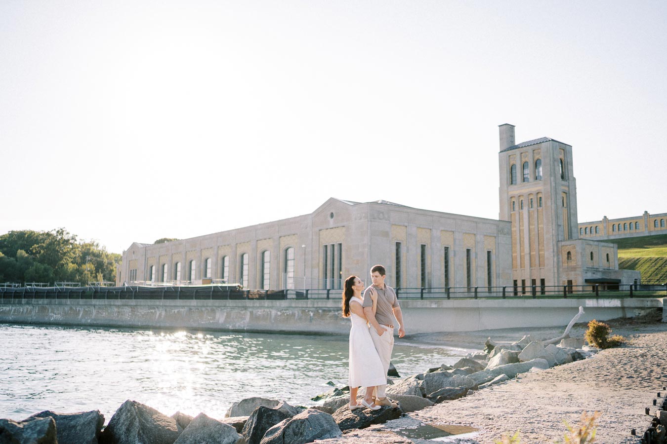 A couple standing on rocks by a calm waterfront, with a historic, industrial-style building and tower in the background under a clear sky.