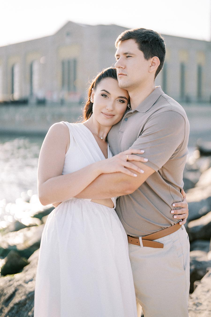 A couple embracing by the waterfront, with soft sunlight illuminating their serene expressions and a historic building in the background, conveying a sense of romance and tranquility.