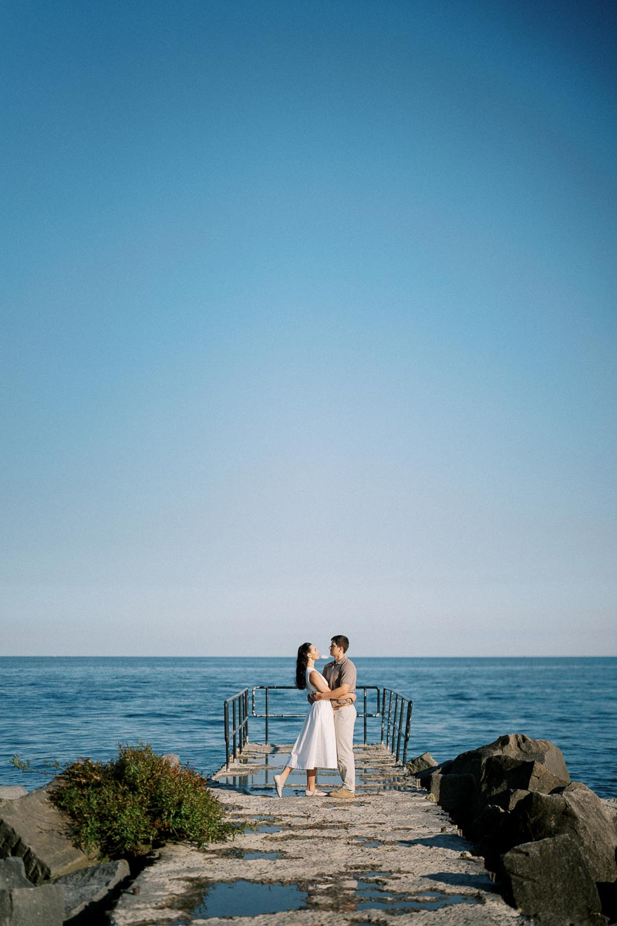 A couple embracing on a rocky pier with a clear blue sky and calm ocean in the background.