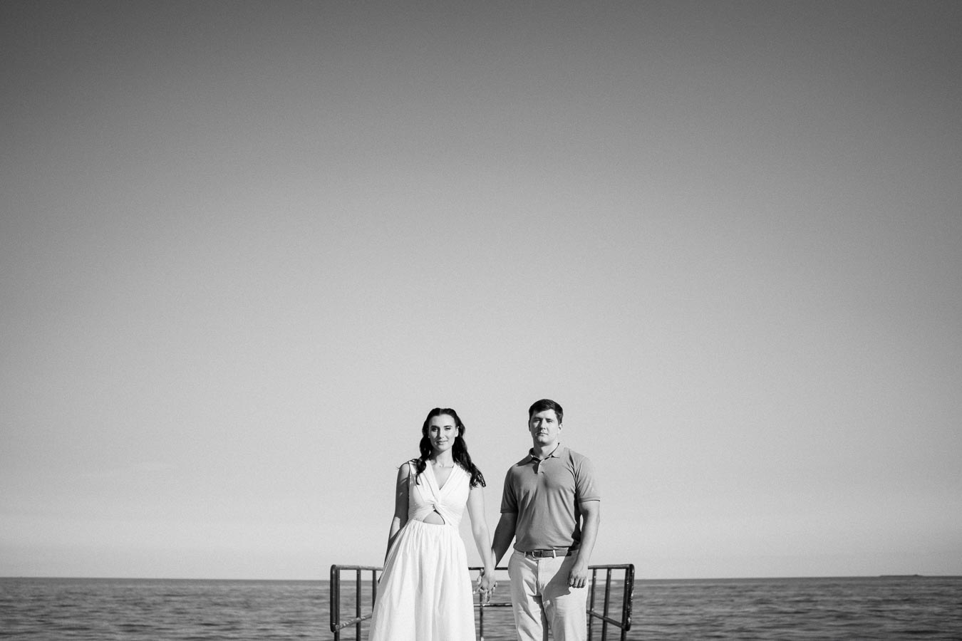 Black and white photo of a couple standing on a dock by the sea, with the horizon visible in the background. The woman is wearing a long white dress and the man is in a polo shirt and pants, holding hands under a clear sky.