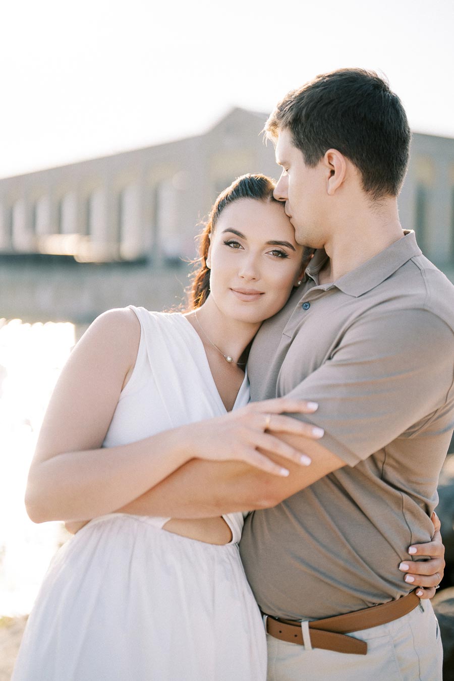 A couple embraces affectionately near a waterfront, with a bridge in the background under a clear sky.