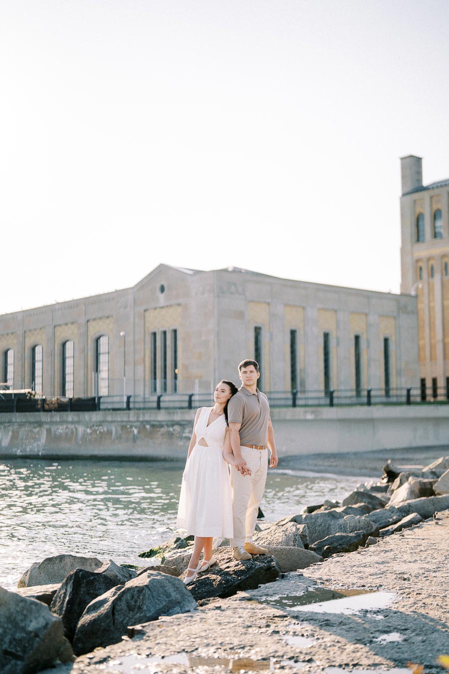Romantic couple posing by the waterfront, standing on rocks in front of an industrial-style building, under a clear sky.