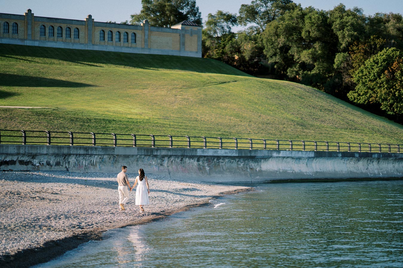 A couple holding hands and walking along a peaceful beach, with gentle waves lapping at the shore, surrounded by lush greenery and a historic building in the background.