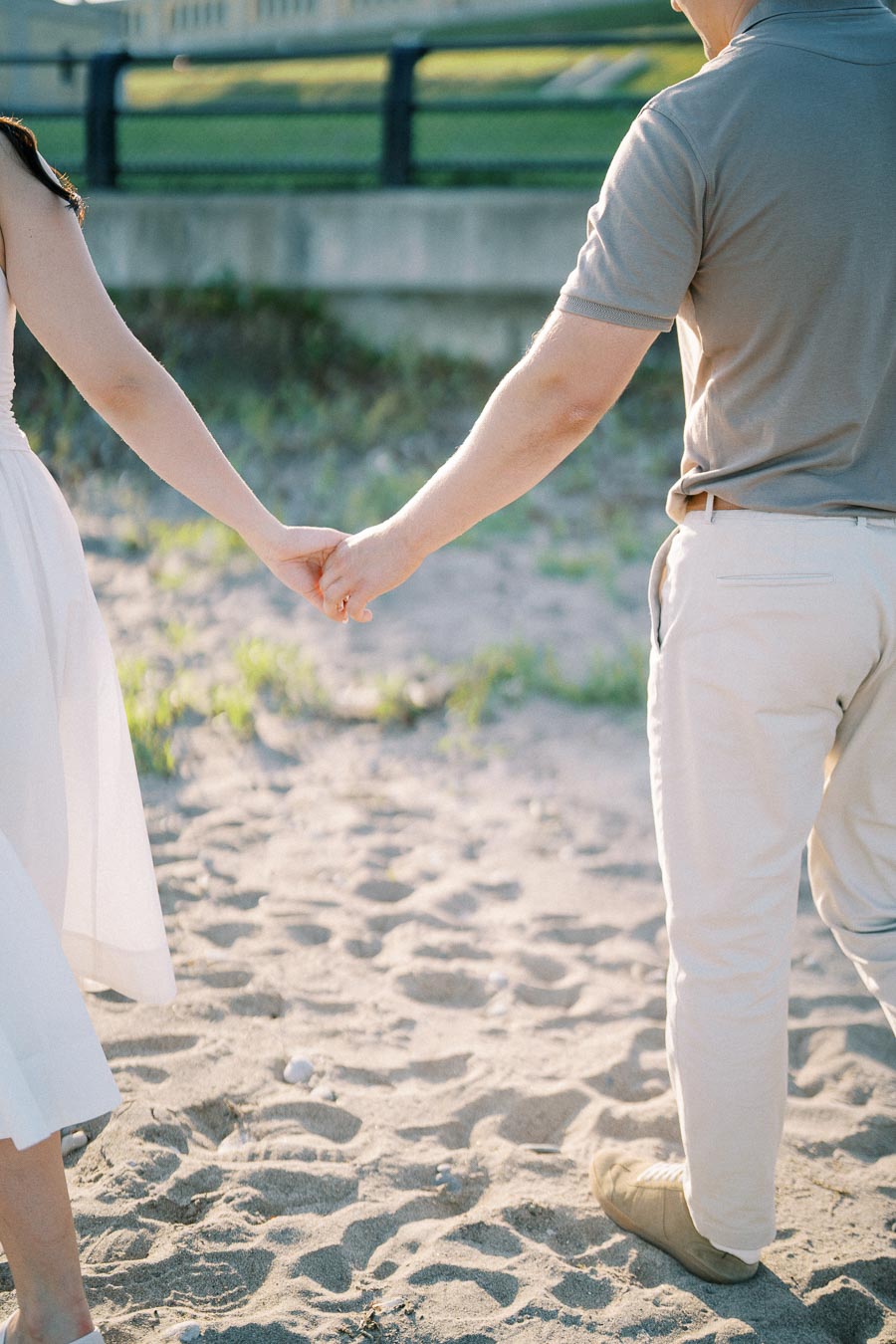 Couple holding hands walking on a sandy beach, wearing casual summer clothing with a blurred grassy background.