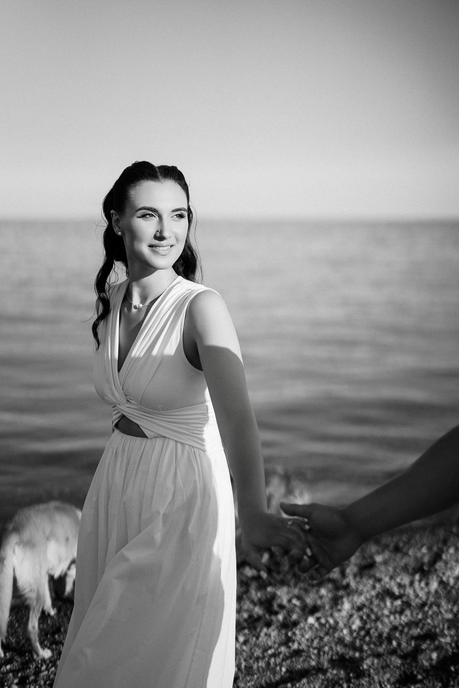 Black and white photo of a woman in a white dress holding hands on a rocky beach with the ocean in the background.