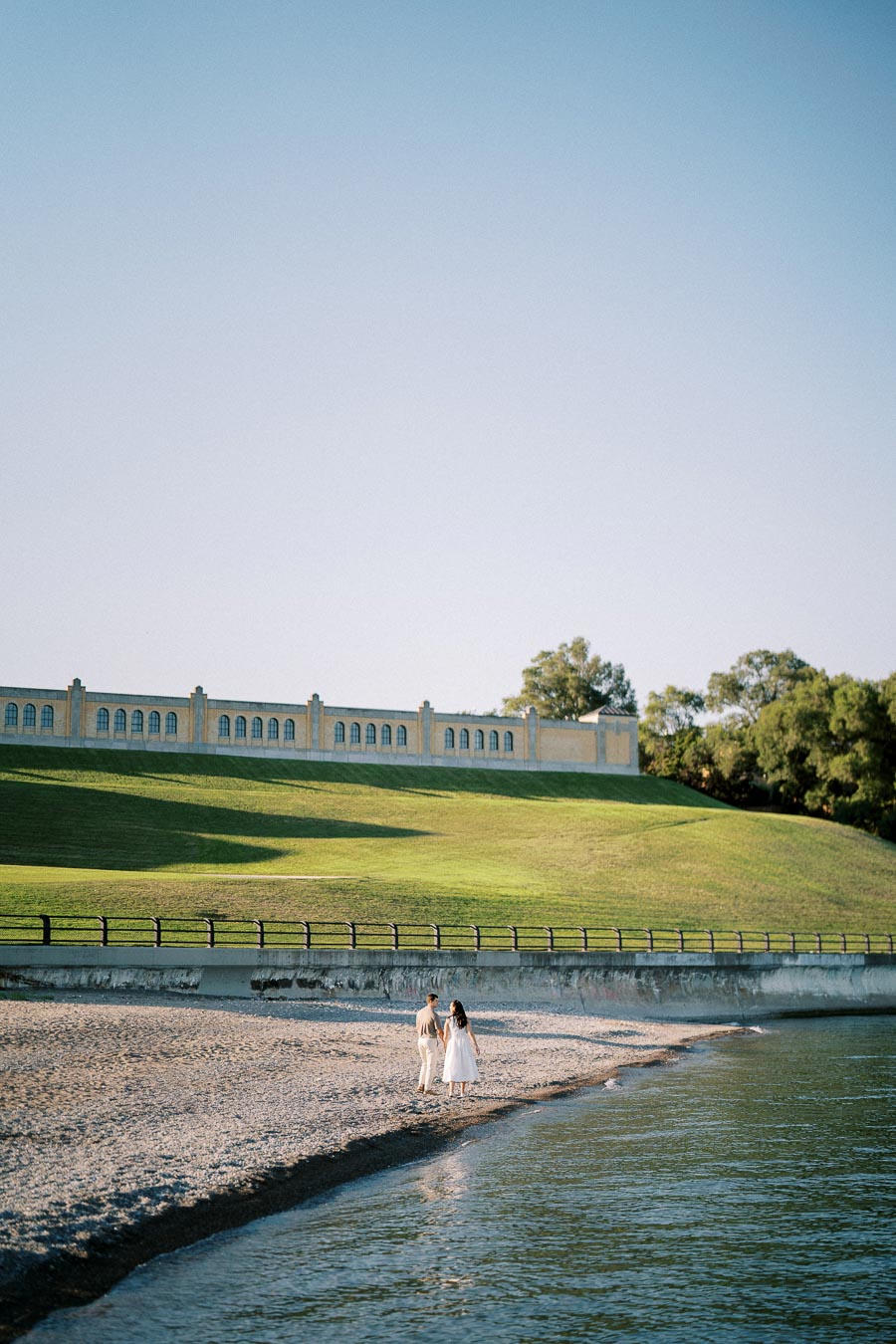 A couple walking along a tranquil lakeside beach with a backdrop of a large historic building and lush green hill under a clear blue sky.