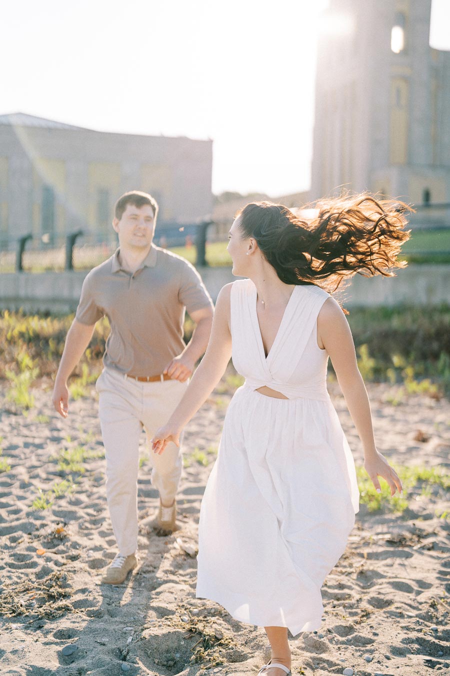 A couple playfully running together on a sunny beach, with the woman in a flowing white dress and the man in a beige shirt and pants, enjoying a romantic day outdoors.