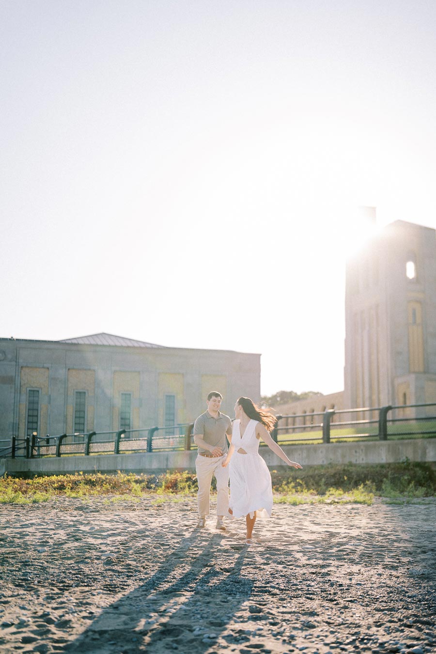 A couple walking on a sunlit beach in front of a historic building, under a clear blue sky.