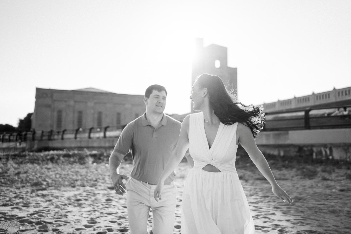 A couple joyfully running on the beach near a historic building during sunset, captured in black and white.