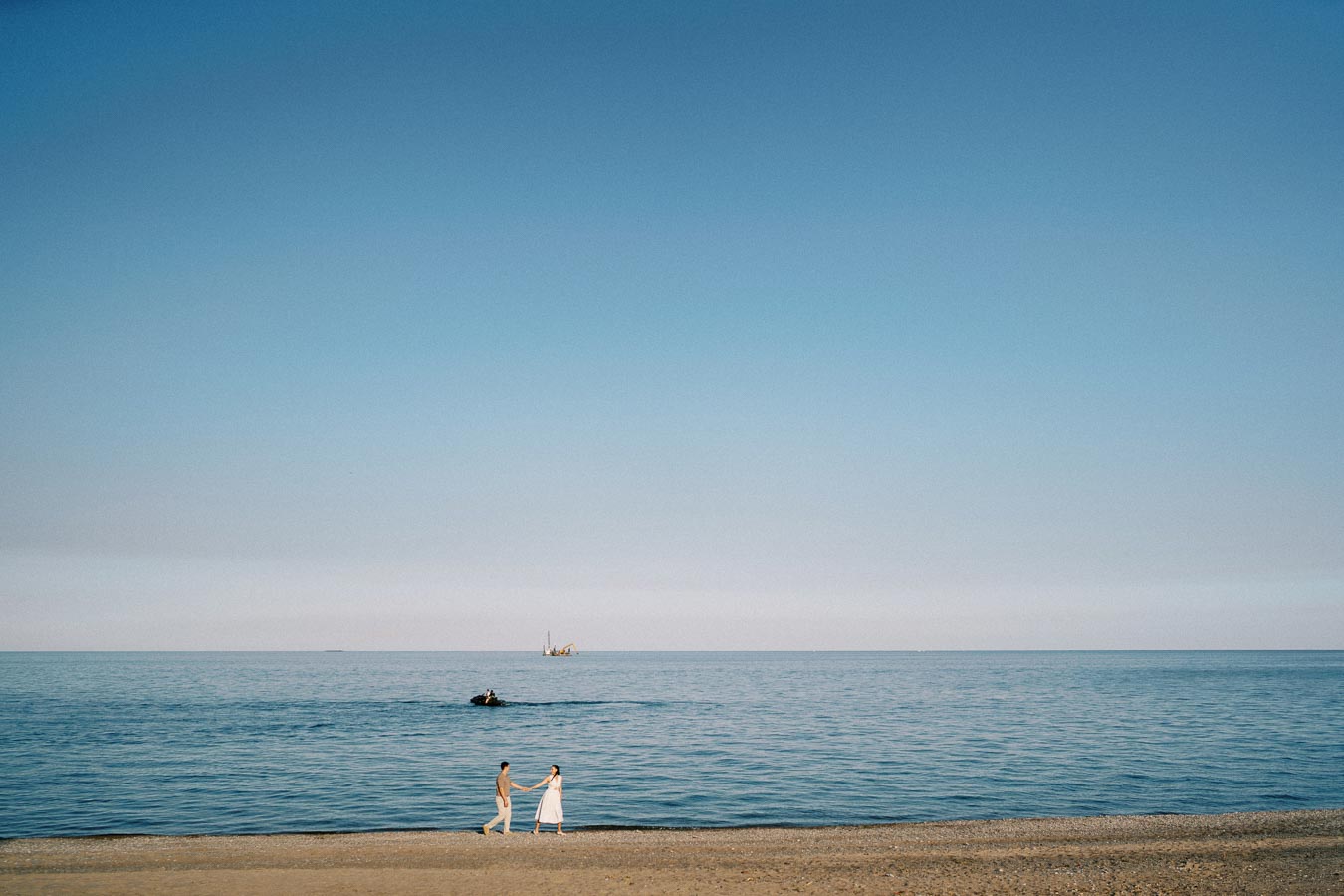 A couple holding hands walking along a serene sandy beach with a calm blue sea and clear sky in the background.