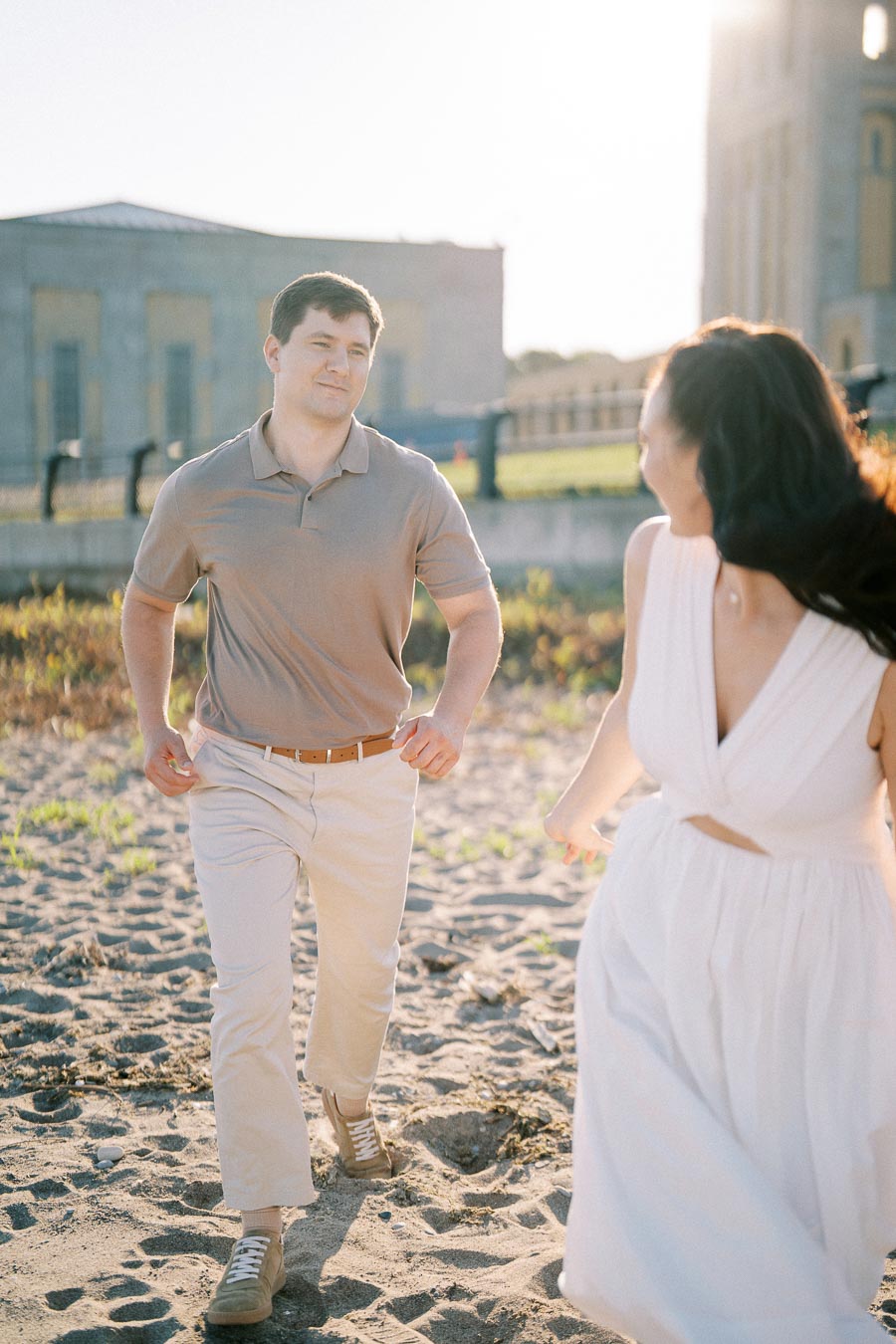 A couple enjoying a sunny day at the beach, with a man in a beige polo shirt and khaki pants smiling at a woman in a white dress, with a historic building in the background.