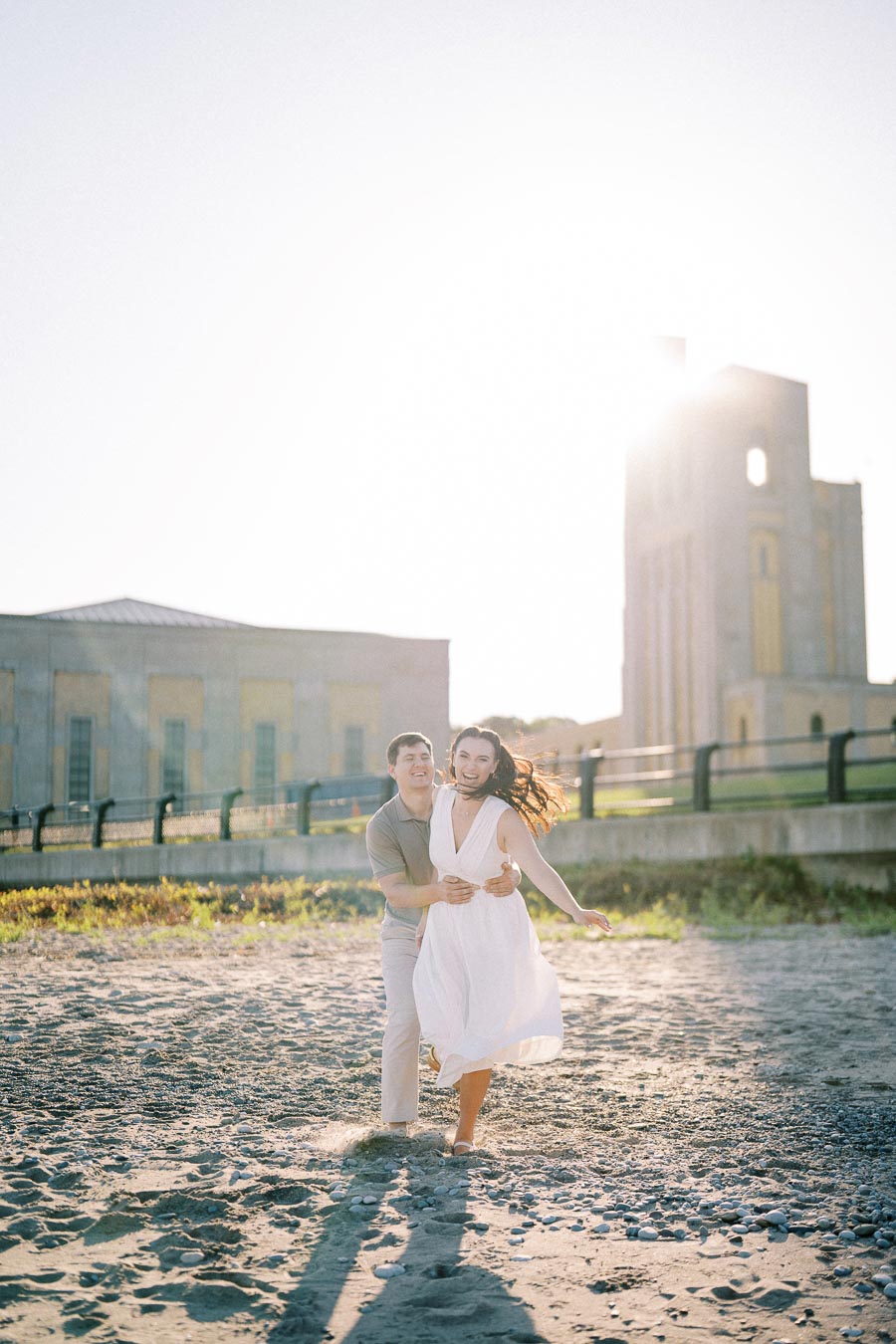 A couple joyfully running on a sandy beach near an old building, with the sun shining brightly in the background.
