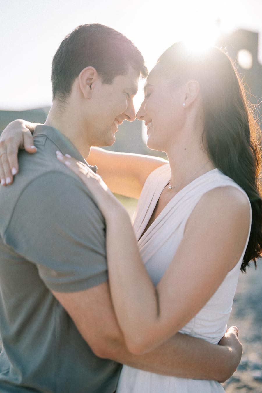 Romantic couple embracing outdoors with sunlight in background, woman in white dress and man in grey shirt, capturing a tender moment.