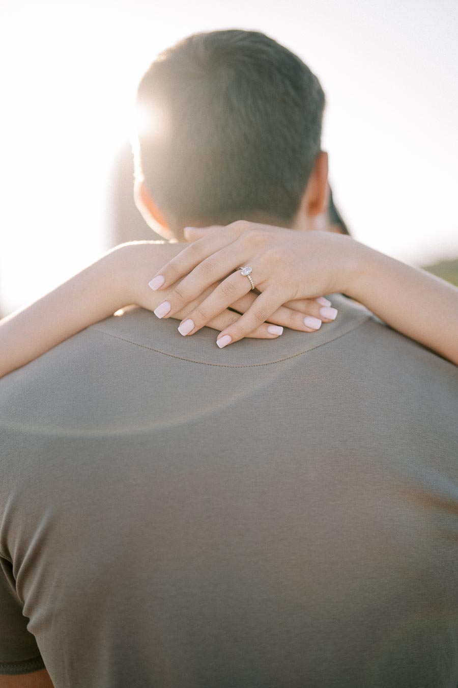 A woman with an elegant engagement ring hugs her partner from behind, her manicured nails gently resting on his shoulder, with sunlight softly illuminating the scene.