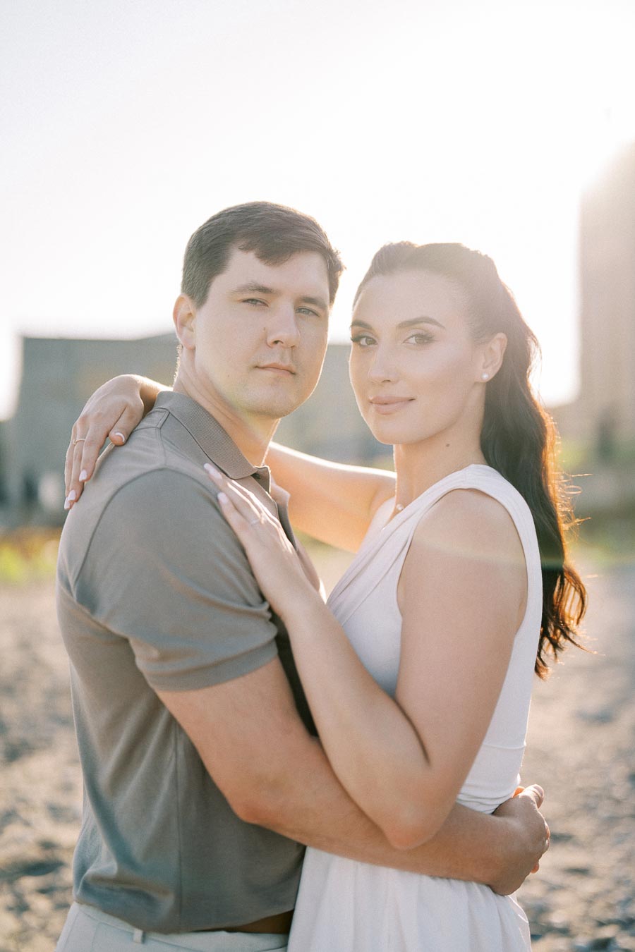 Couple embracing on a sunny beach, looking at the camera, with a soft glow highlighting them.