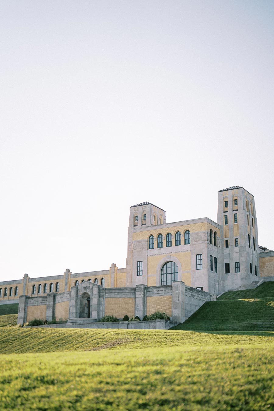 Historic stone building with towers and large windows, set on a grassy hill under a clear sky.