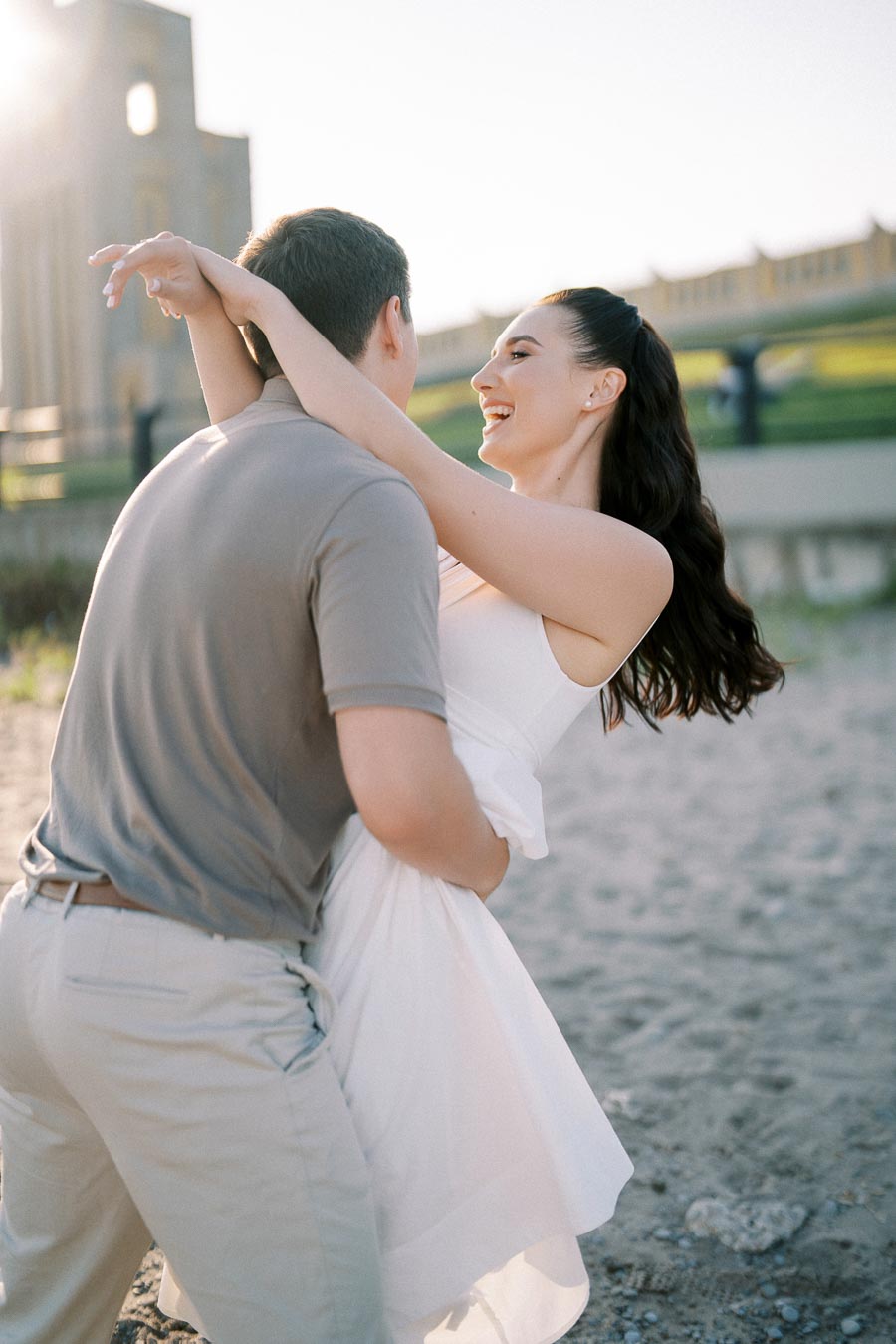 A couple dancing joyfully on a sunlit beach, with the woman smiling as she embraces her partner in a casual summer setting.