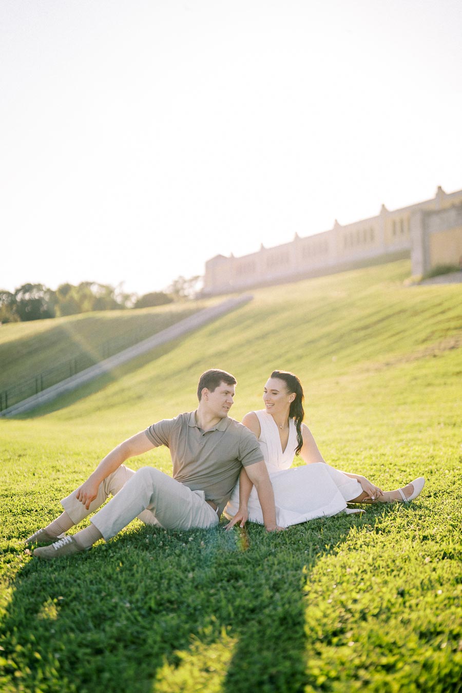 A couple sitting on the grass in a sunlit park, engaging in a pleasant conversation with a historic building in the background.