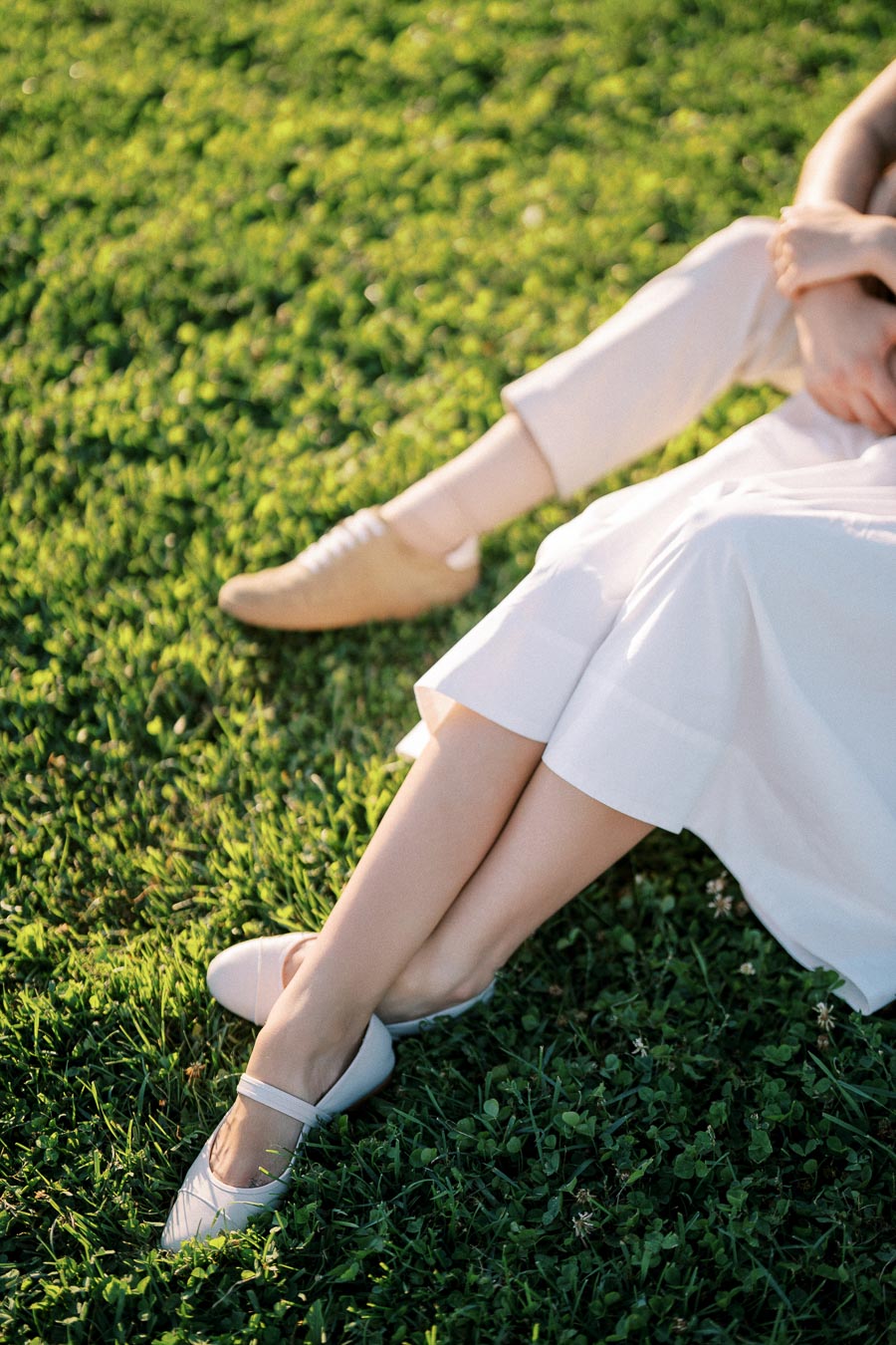 Two people sitting on grass, showing crossed legs with one wearing beige shoes and light pants, and the other wearing white ballet flats and a flowing white skirt, in a sunlit outdoor setting.