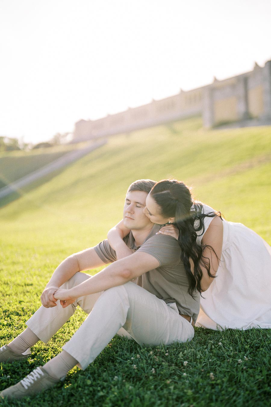 A woman in a white dress lovingly embraces a man in casual attire as they sit on green grass with a slight hill and a building in the background during a sunny day.