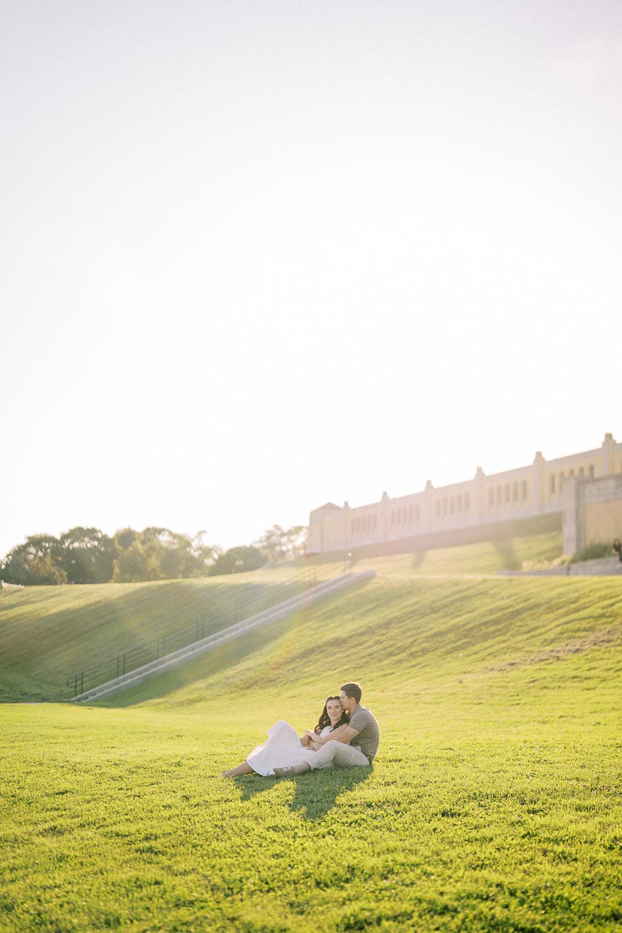A couple sitting on a grassy hill under a sunny sky, with a historic building in the background, enjoying a romantic moment outdoors.