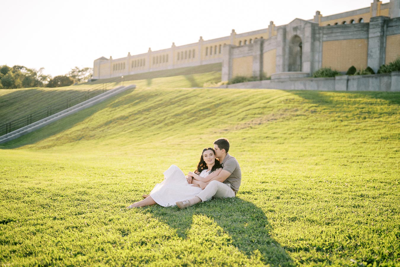 A couple sitting together on a grassy hill in a park, with a large building in the background and warm sunlight casting long shadows.