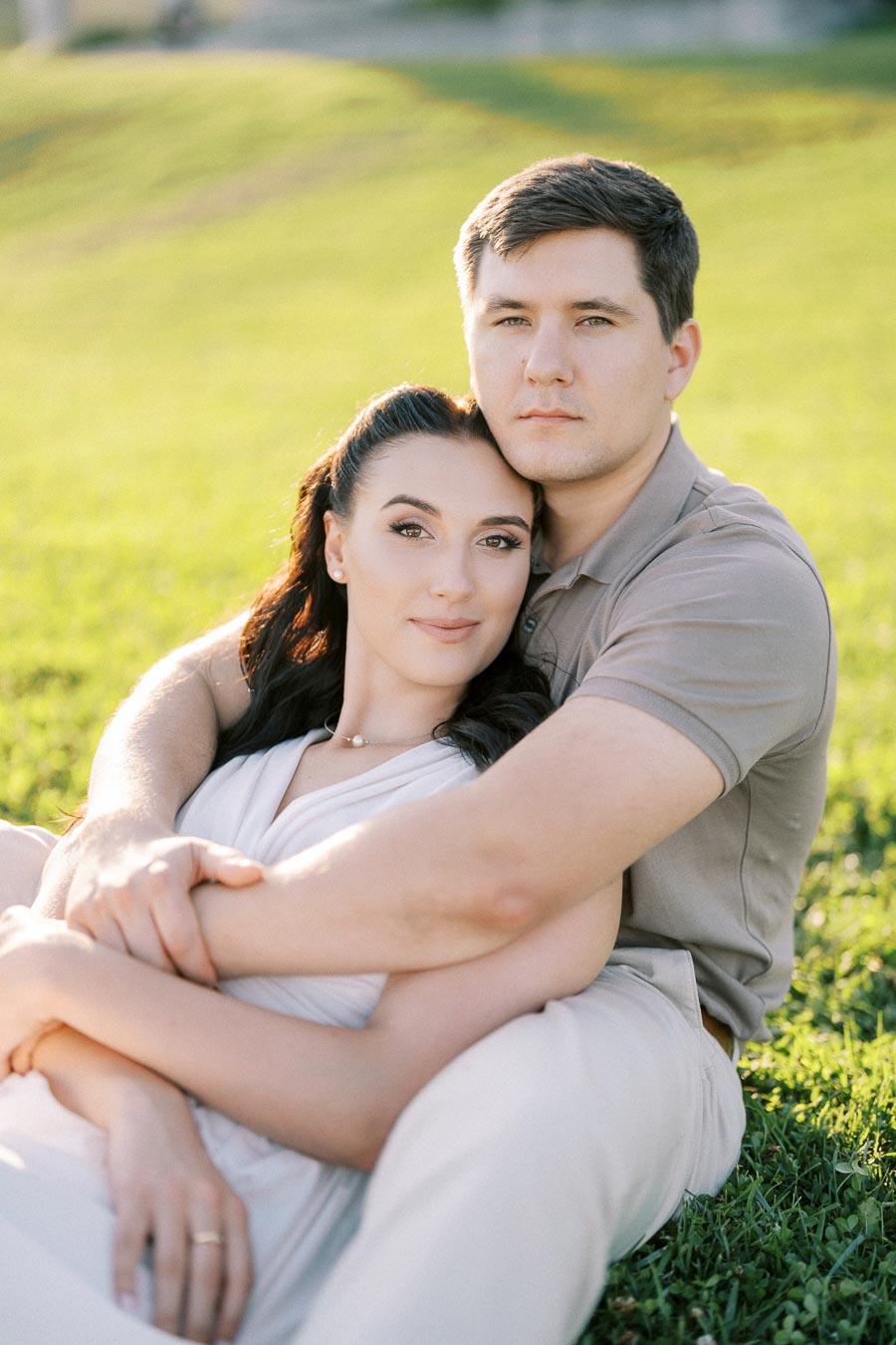 A couple sitting on grass in a park, embraced and smiling warmly at the camera, with sunlight creating a serene and romantic atmosphere.