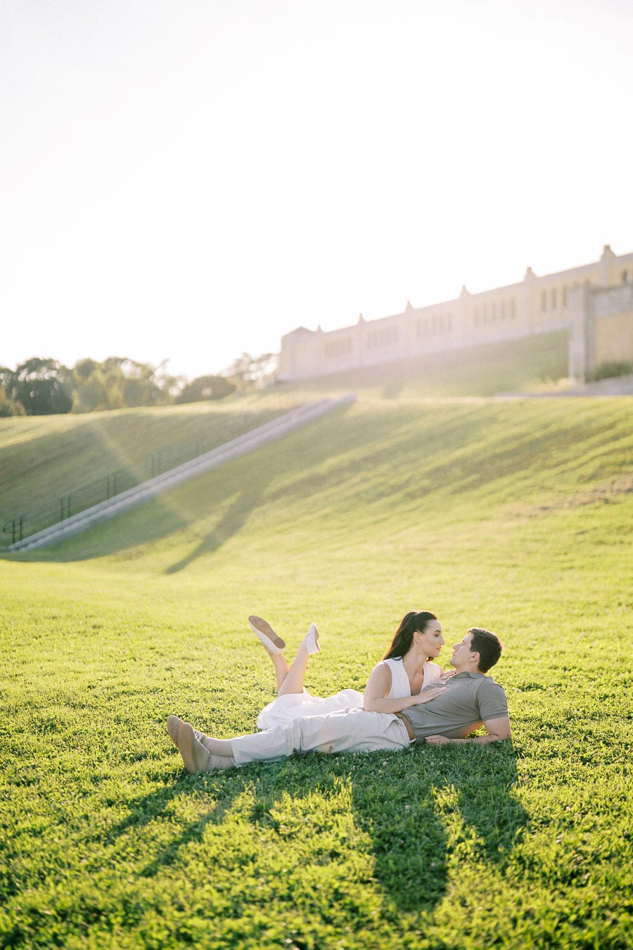 A couple lying on a grassy hill, embracing under sunny skies, with a historic building in the background.