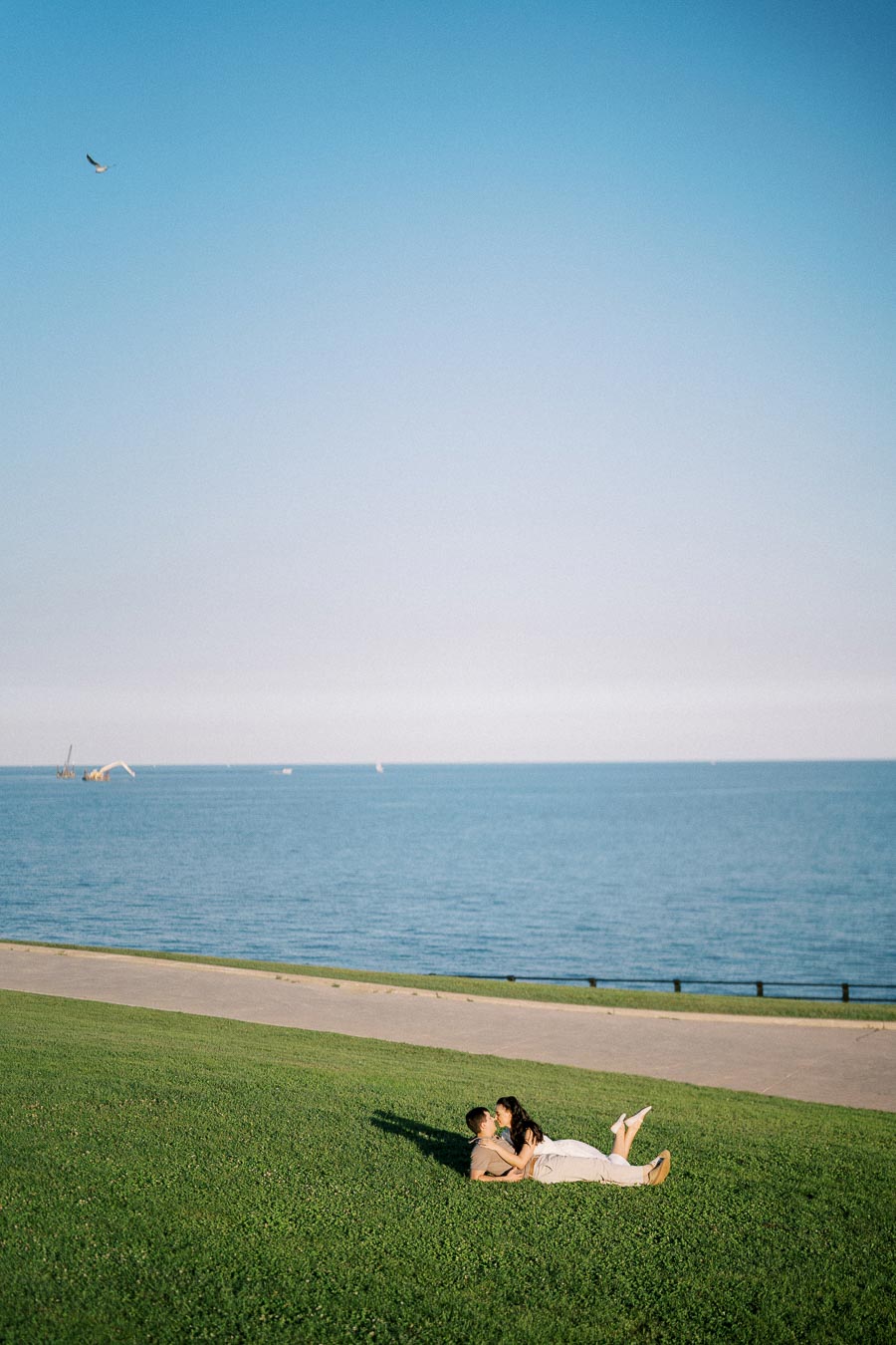 A couple lying on a grassy hill overlooking a serene blue lake under a clear sky.