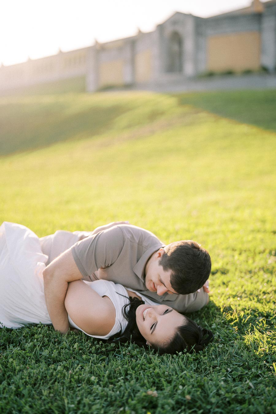 A couple lying on a grassy field, embracing and smiling warmly at each other, with a soft-focus background showcasing a stone building in the distance under natural sunlight.