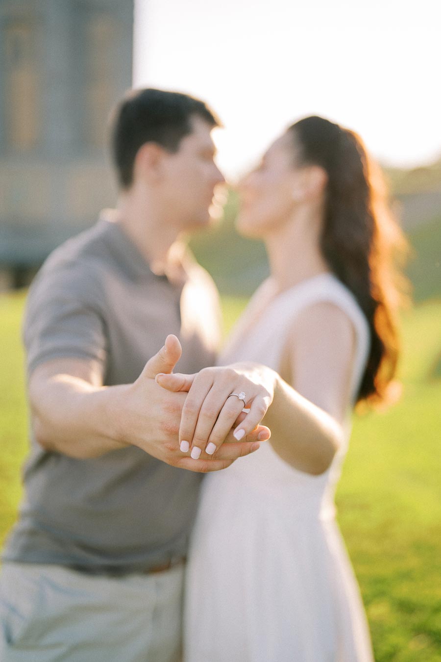 A couple holding hands with a focus on an engagement ring in a sunlit outdoor setting, symbolizing love and commitment.