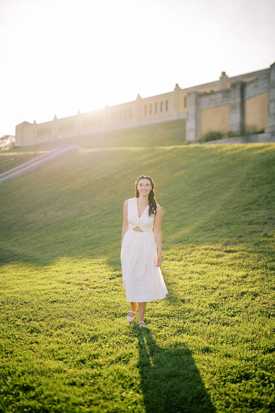 A woman in a white dress walks on a sunlit grassy hill with a historic building in the background.
