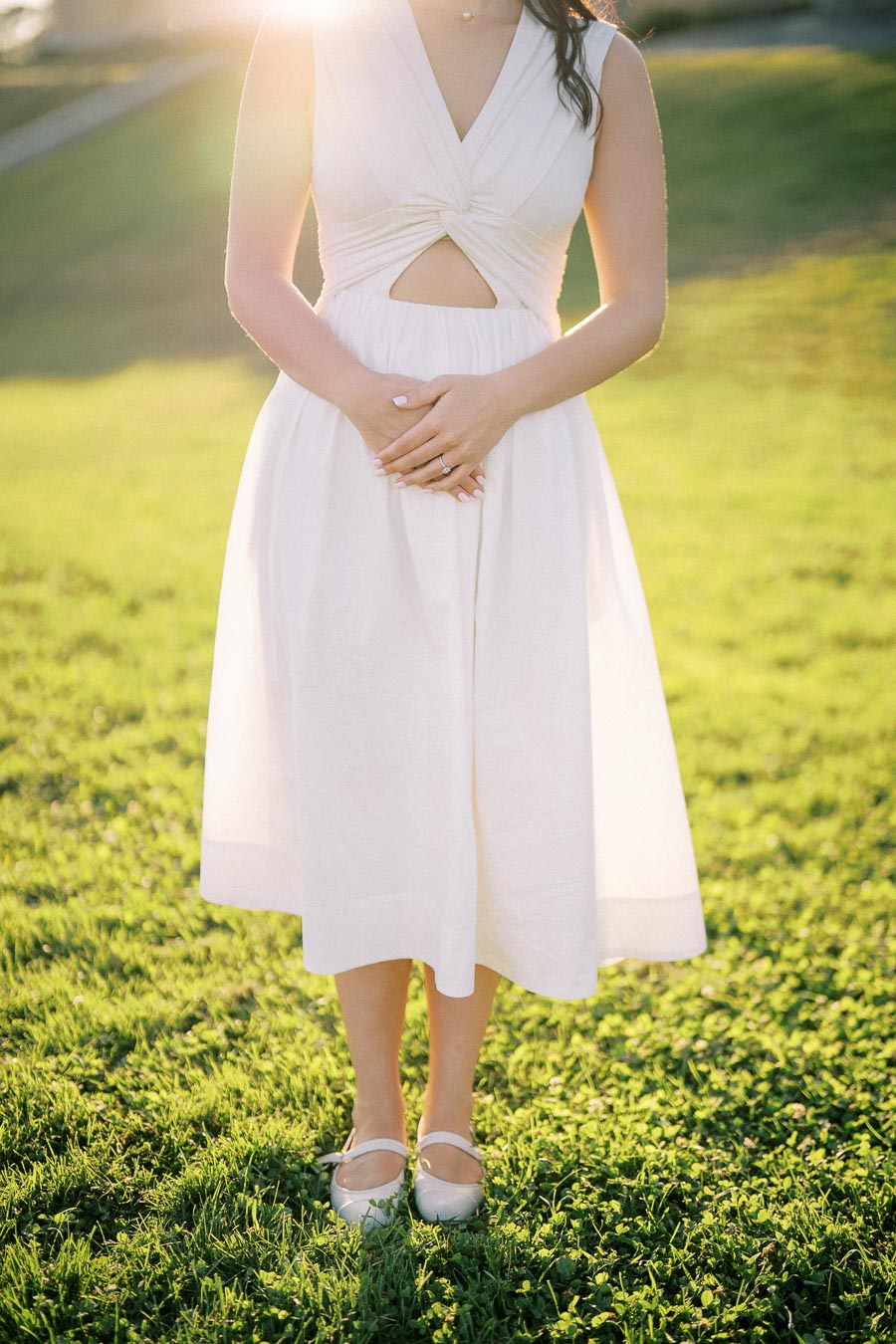 A woman in a white sleeveless dress stands on green grass, bathed in sunlight.
