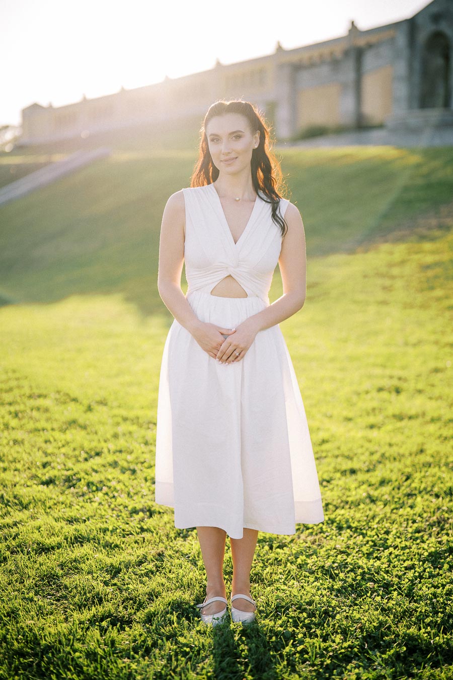 A woman in a white dress standing on a grassy field with a historic building in the background, illuminated by soft sunlight.