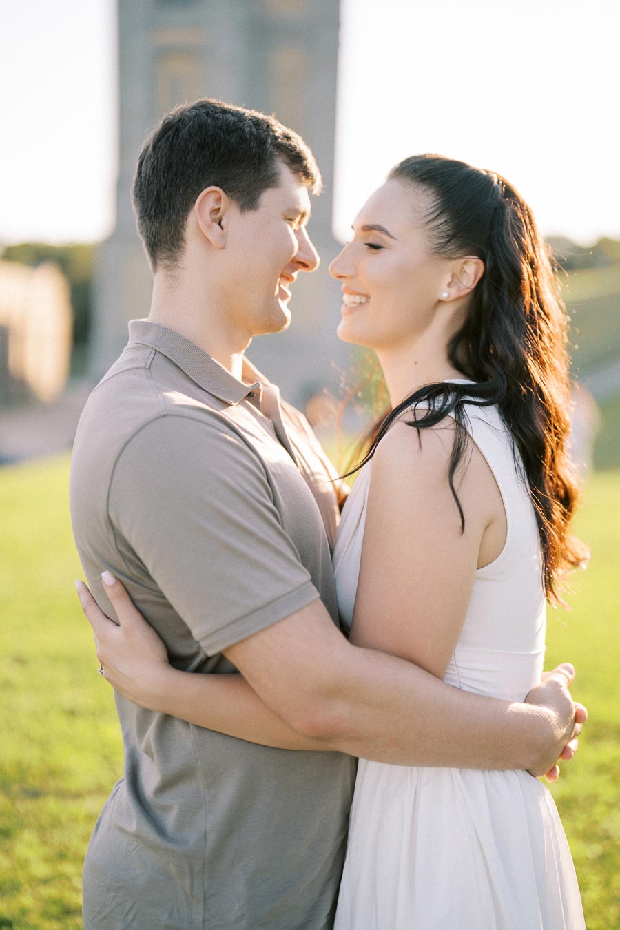 A couple embracing in a sunlit park with a historic monument in the background, capturing a romantic and joyful moment outdoors.