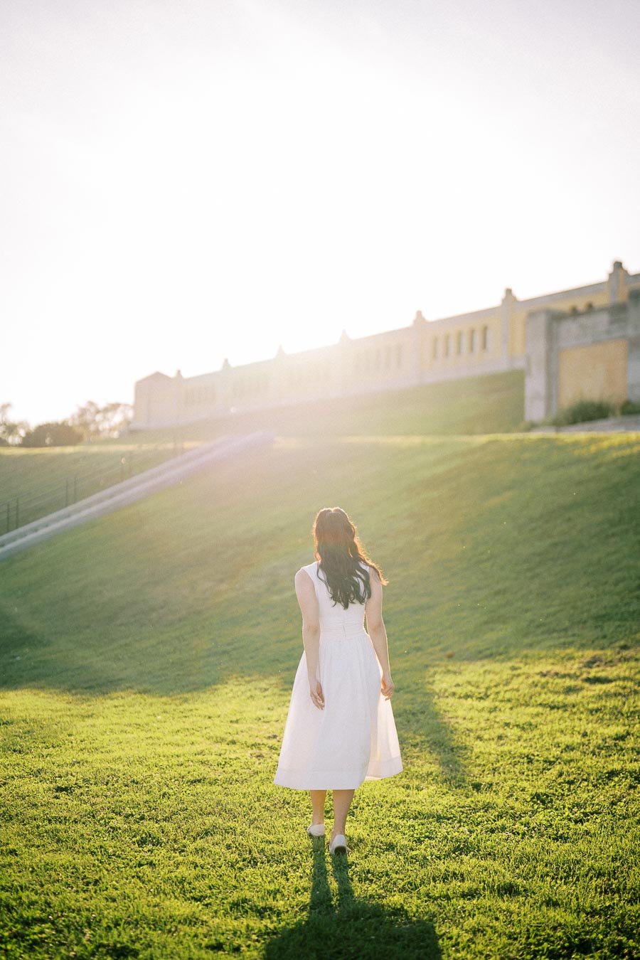 A woman in a white dress walks up a grassy hill towards a large yellow building under bright sunlight.