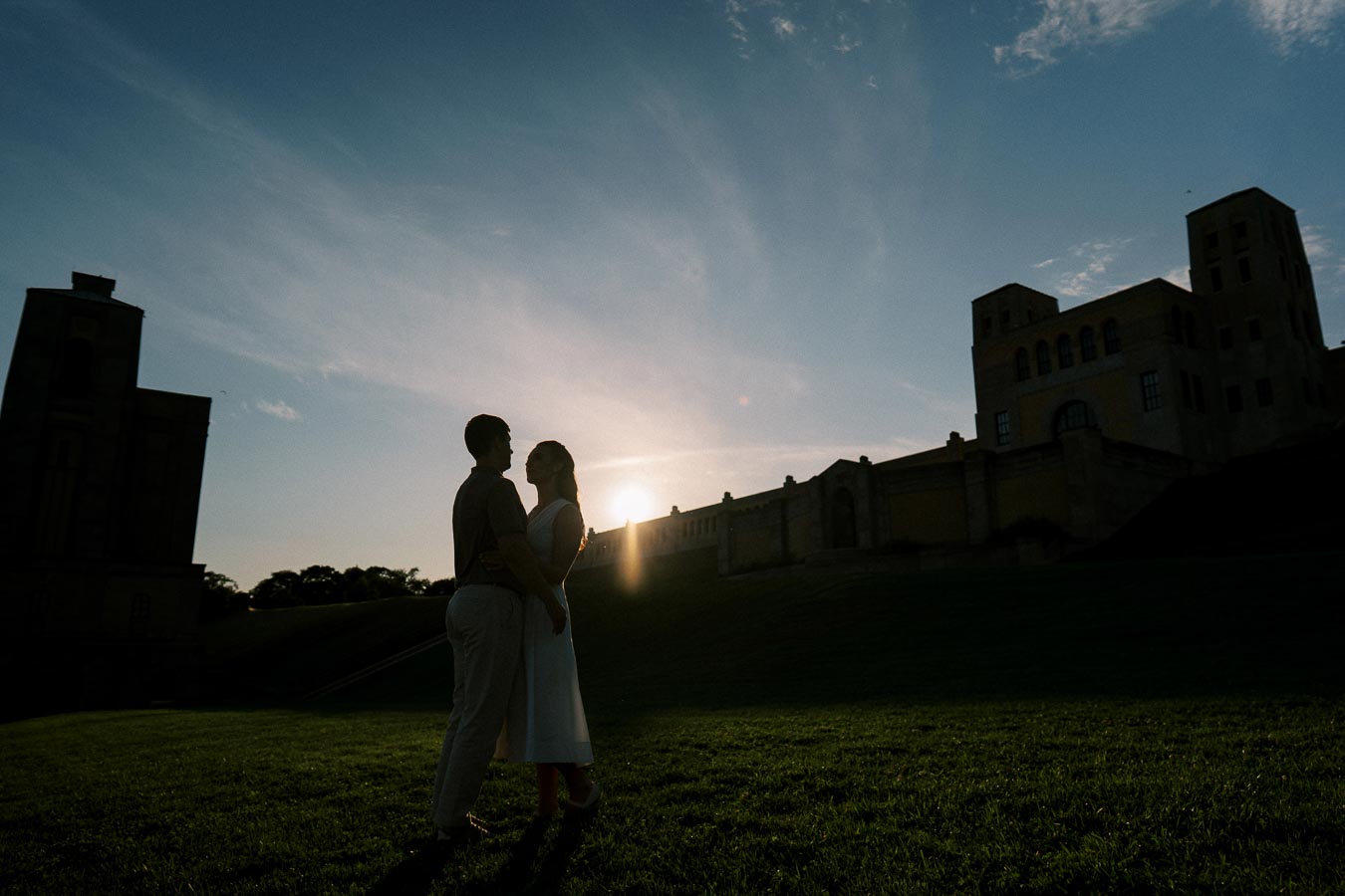 A romantic couple silhouetted against a scenic sunset, embraced on a grassy hill with historic buildings in the background.