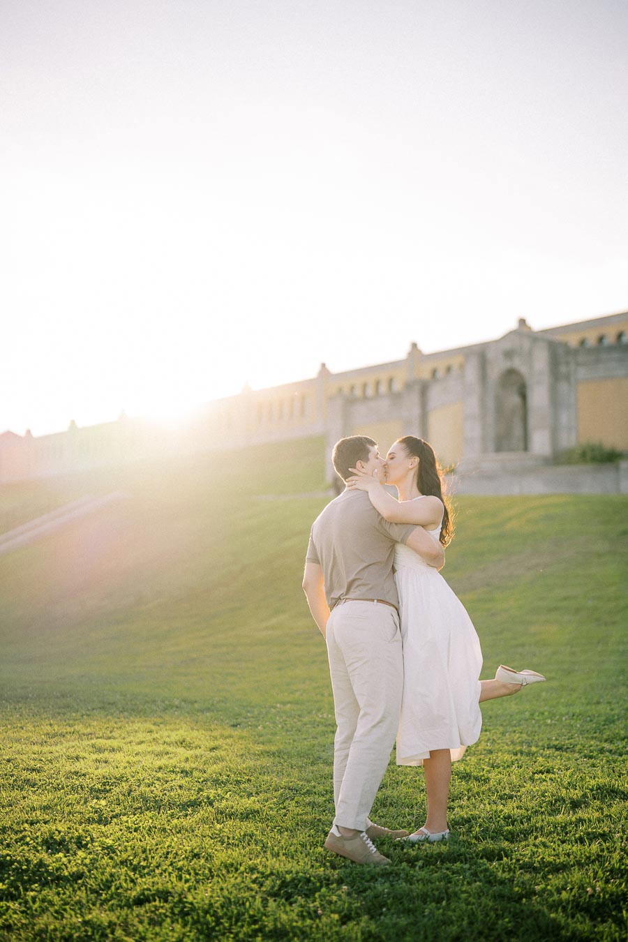 A couple embraces and kisses on a sunlit grassy field, with a historic stone building in the background, creating a romantic and serene atmosphere.