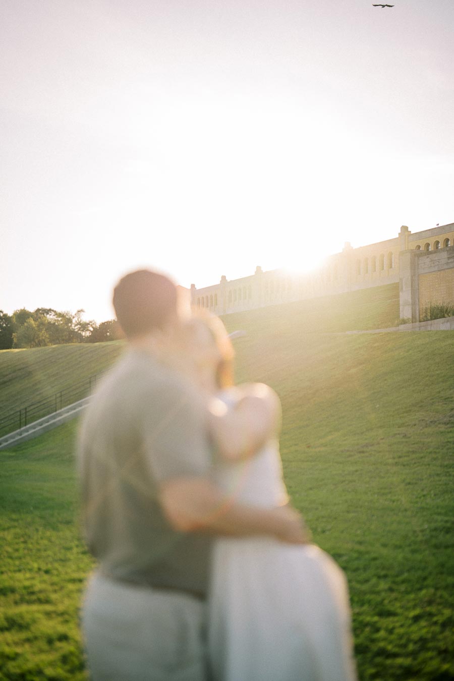 A couple embraces in a grassy field with the sun setting behind a historic building, creating a warm and romantic atmosphere.