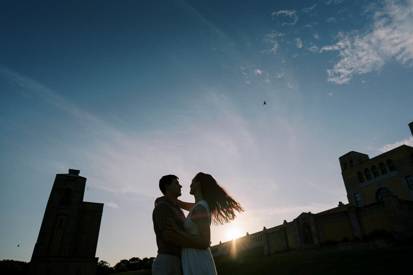 Couple embracing during sunset at a park with historical buildings in the background.