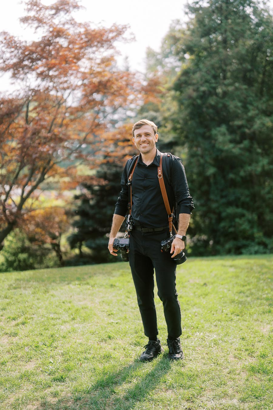 Professional photographer standing in a scenic outdoor setting with lush greenery and autumn trees, wearing camera gear and smiling confidently.