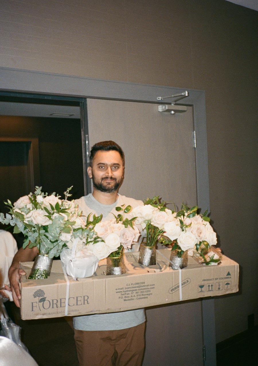 Man carrying a box of fresh white roses in vases, labeled Florecer from Colombia, inside a room with a gray door.