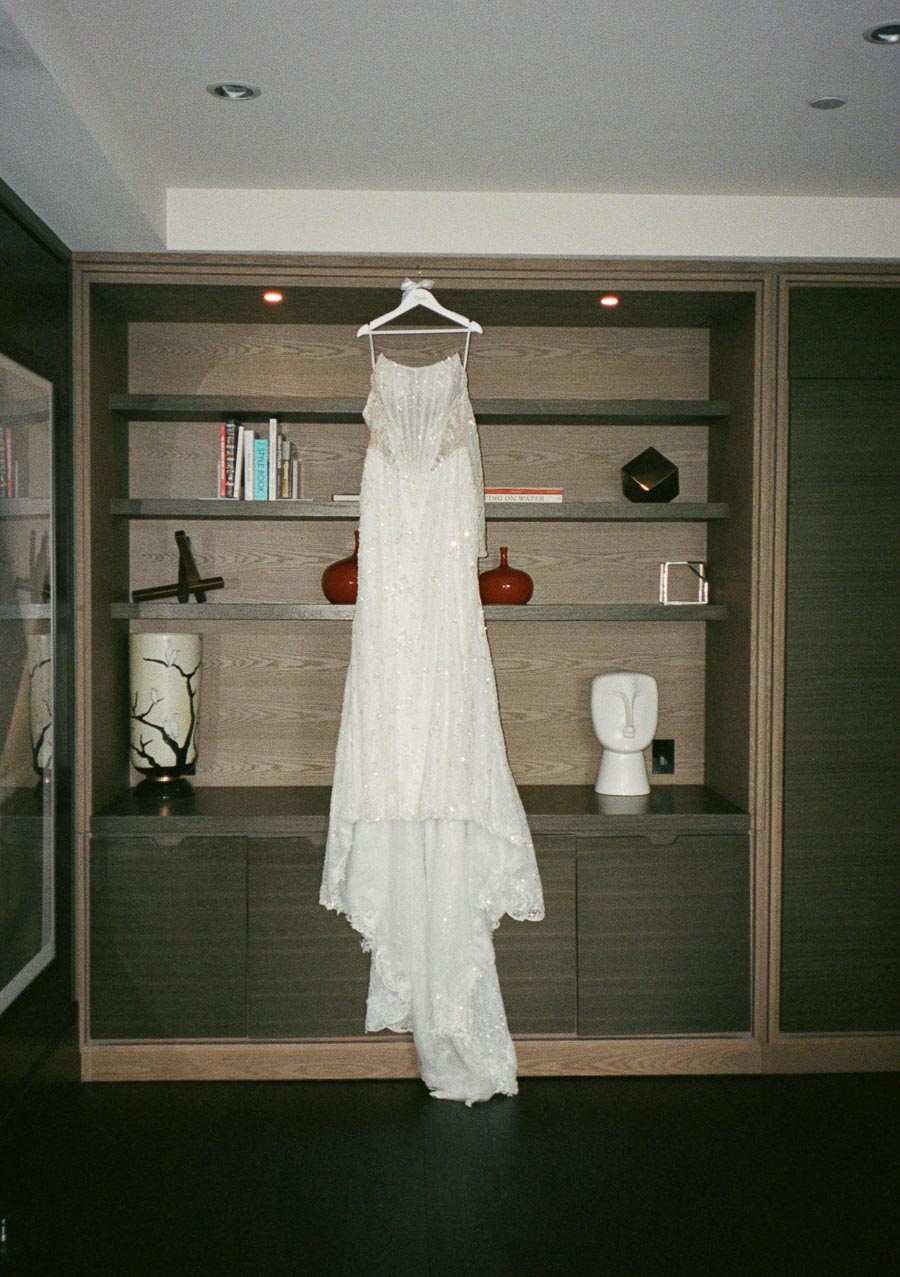 Elegant white wedding dress with intricate beadwork hanging on a wooden bookshelf, surrounded by decorative items and books.