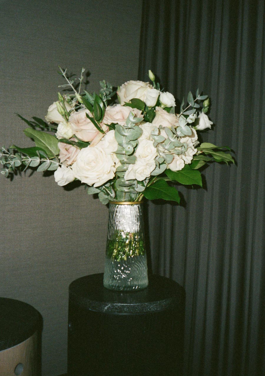 A bouquet of white and pink roses with greenery in a textured glass vase on a dark table against a draped curtain backdrop.