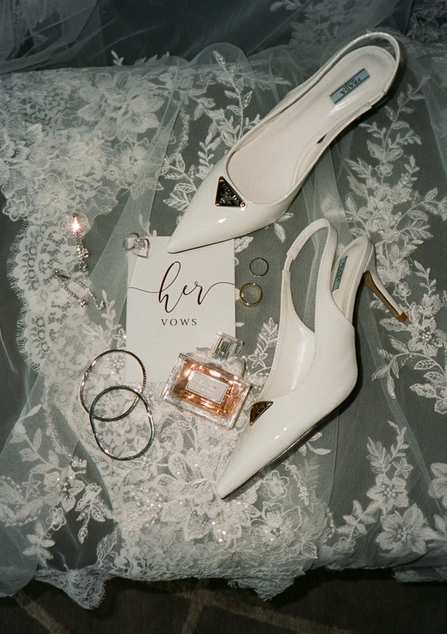 Bridal accessories and details including white high-heeled shoes, a bottle of perfume, wedding vows card, rings, and lace fabric, arranged elegantly.