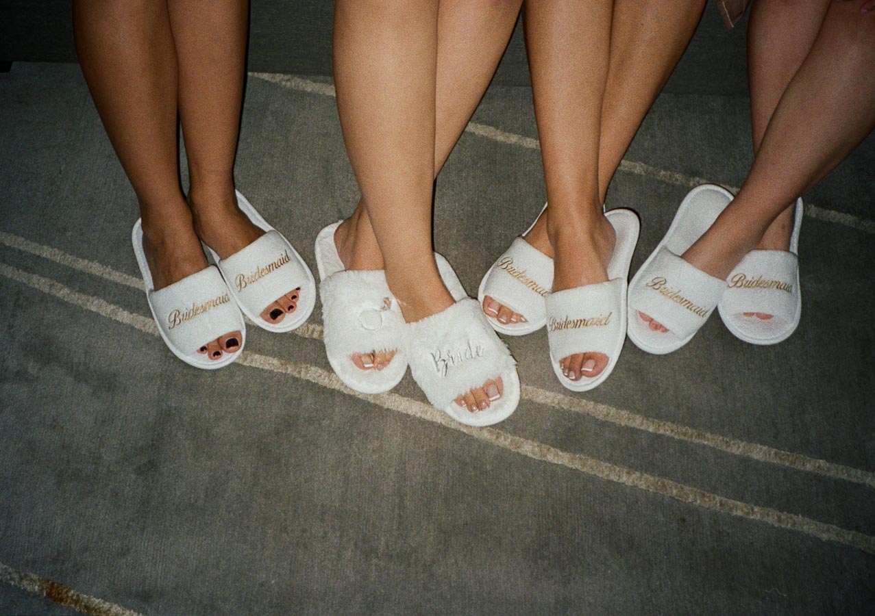 Wedding party in cozy white slippers with Bridesmaid and Bride embroidery, featuring four women with bare legs and painted nails on a gray striped carpet.